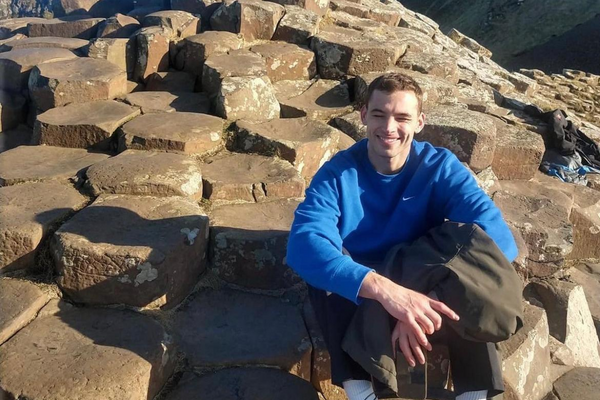 Photo of Isaac Ricca-Righardson,  a pro bono barrister for BiD, sitting outside on some rocks. He is wearing a blue long sleeve shirt and dark trousers. 