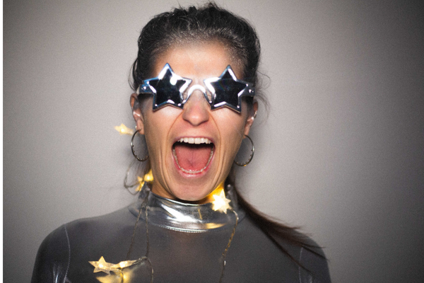 Freya is wearing silver star shaped glasses and a silver disco suit. She has star shaped fairy lights draped over her. She is smiling and facing the camera