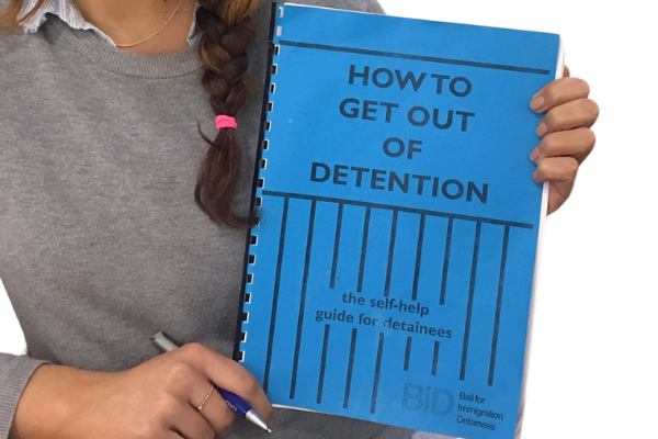 image of a person holding up a blue booklet which says 'how to get out of detention: the self help guide for detainees'